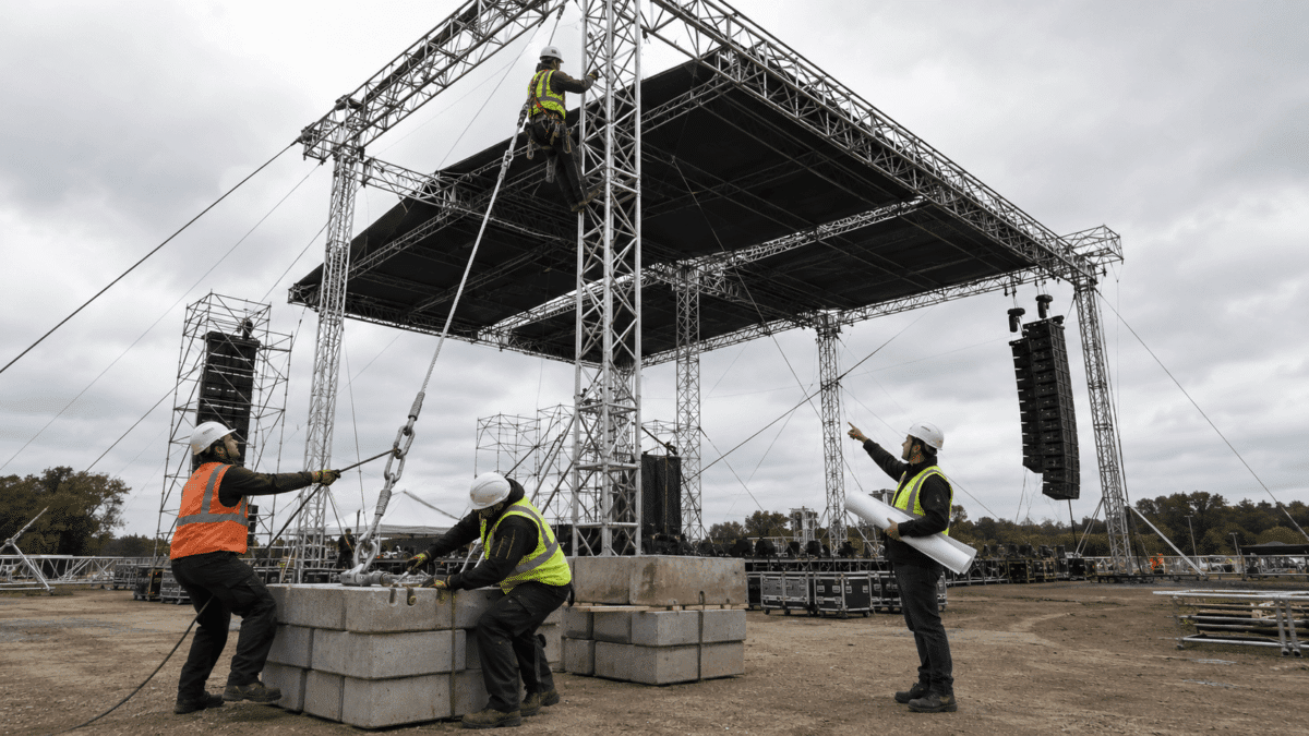 Three construction workers in safety vests and hard hats guide rigging equipment around concrete blocks beneath a large temporary stage structure with steel trusses and overhead lighting rigs.